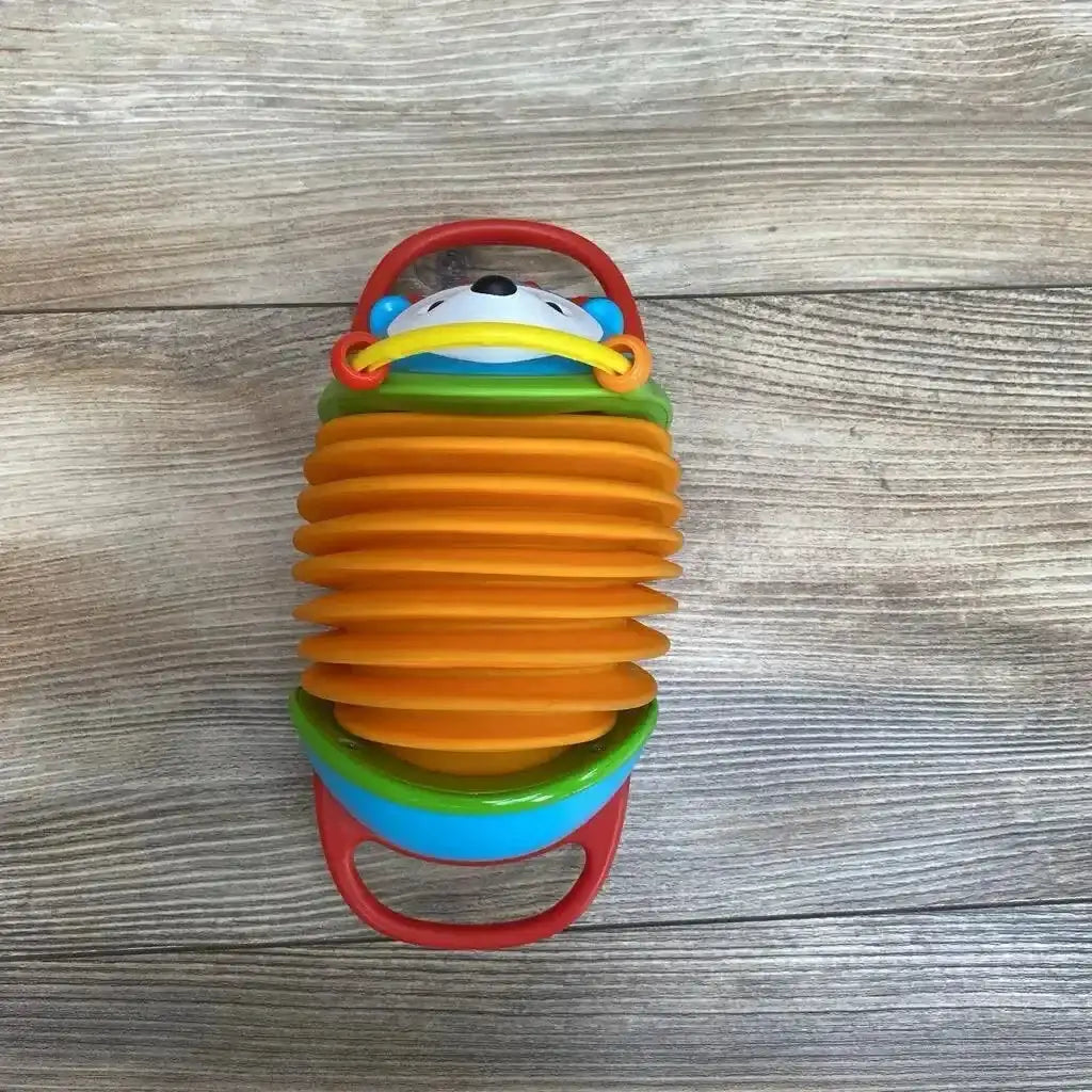 Top-down view of Skip Hop hedgehog accordion toy showcasing orange expandable bellows, red handles, and smiling hedgehog face on wood background