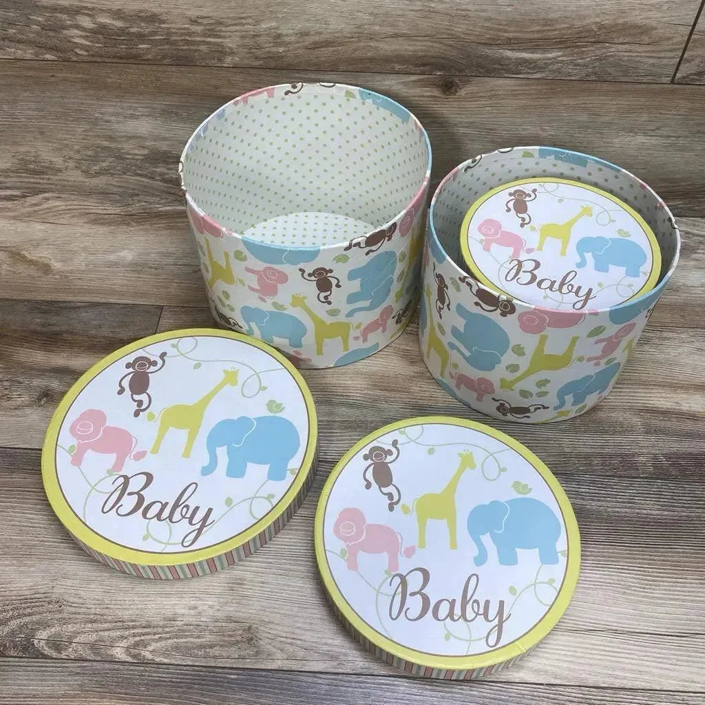 Angled view of a 3-piece set of round nesting baby storage trunks showing pastel elephant, giraffe, and monkey print with 'Baby' lids on wood floor