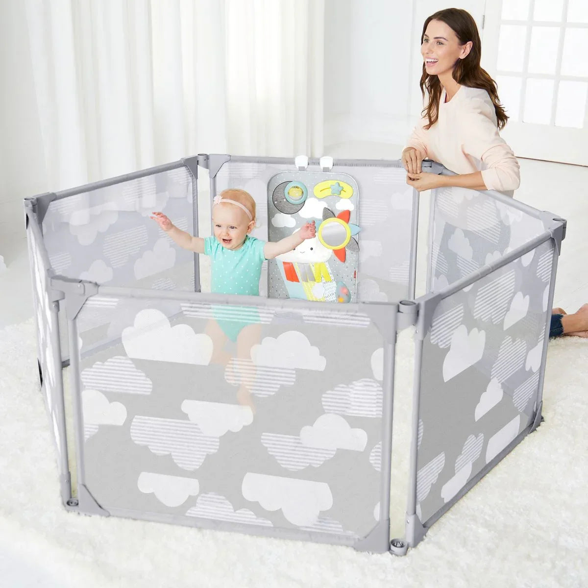 Baby playing in cloud-themed playpen with toys, supervised by woman in bright room