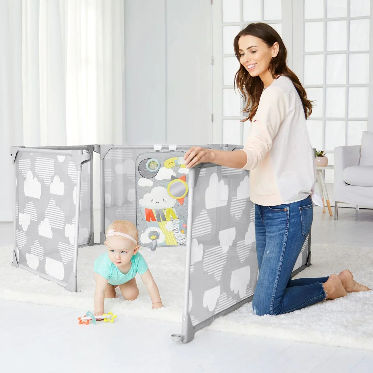 Mom with baby in gray cloud-patterned playpen in bright living room with toys