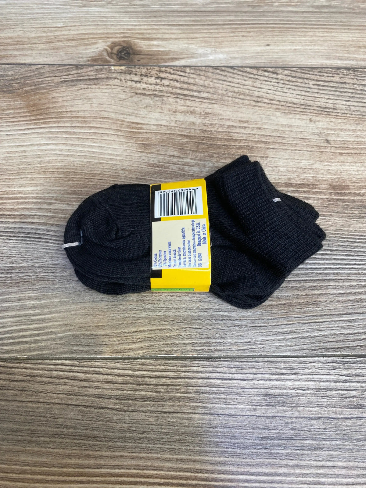 Top down view of three pack black baby socks with yellow label, ribbed cuff and ankle height on wood floor background