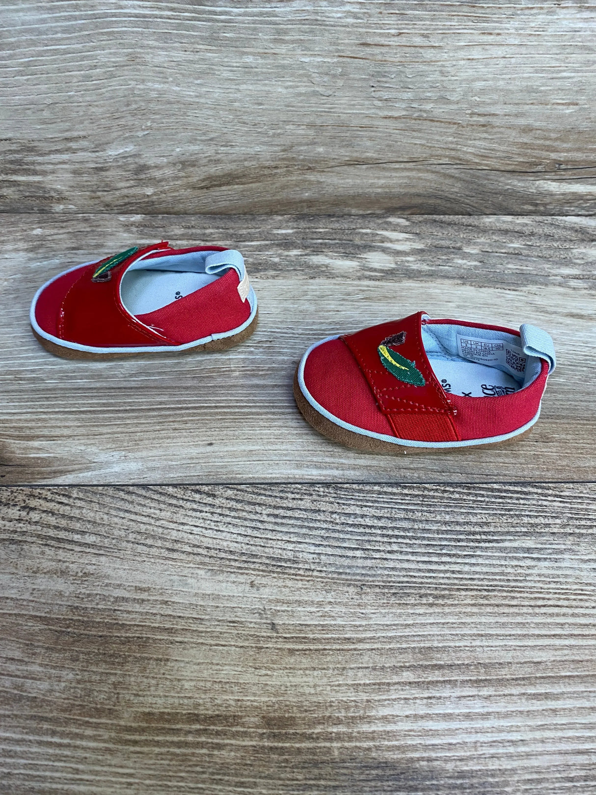 Top-down view of TOMS red crib shoes size 3C with embroidered green banana on canvas, wood background