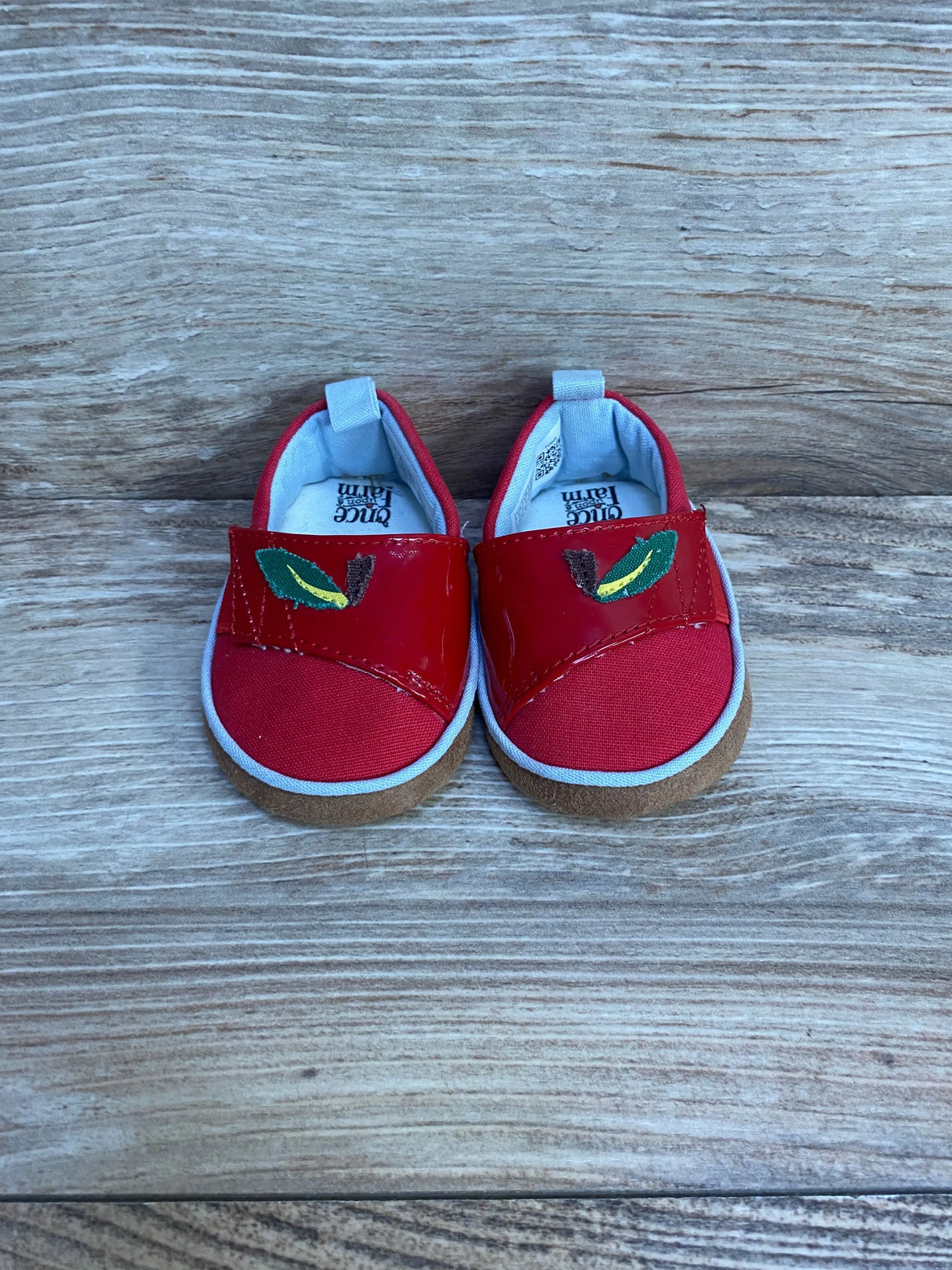 Front view of TOMS Alpargata red crib shoes showcasing patent strap with embroidered green leaf and tan suede sole on wooden background