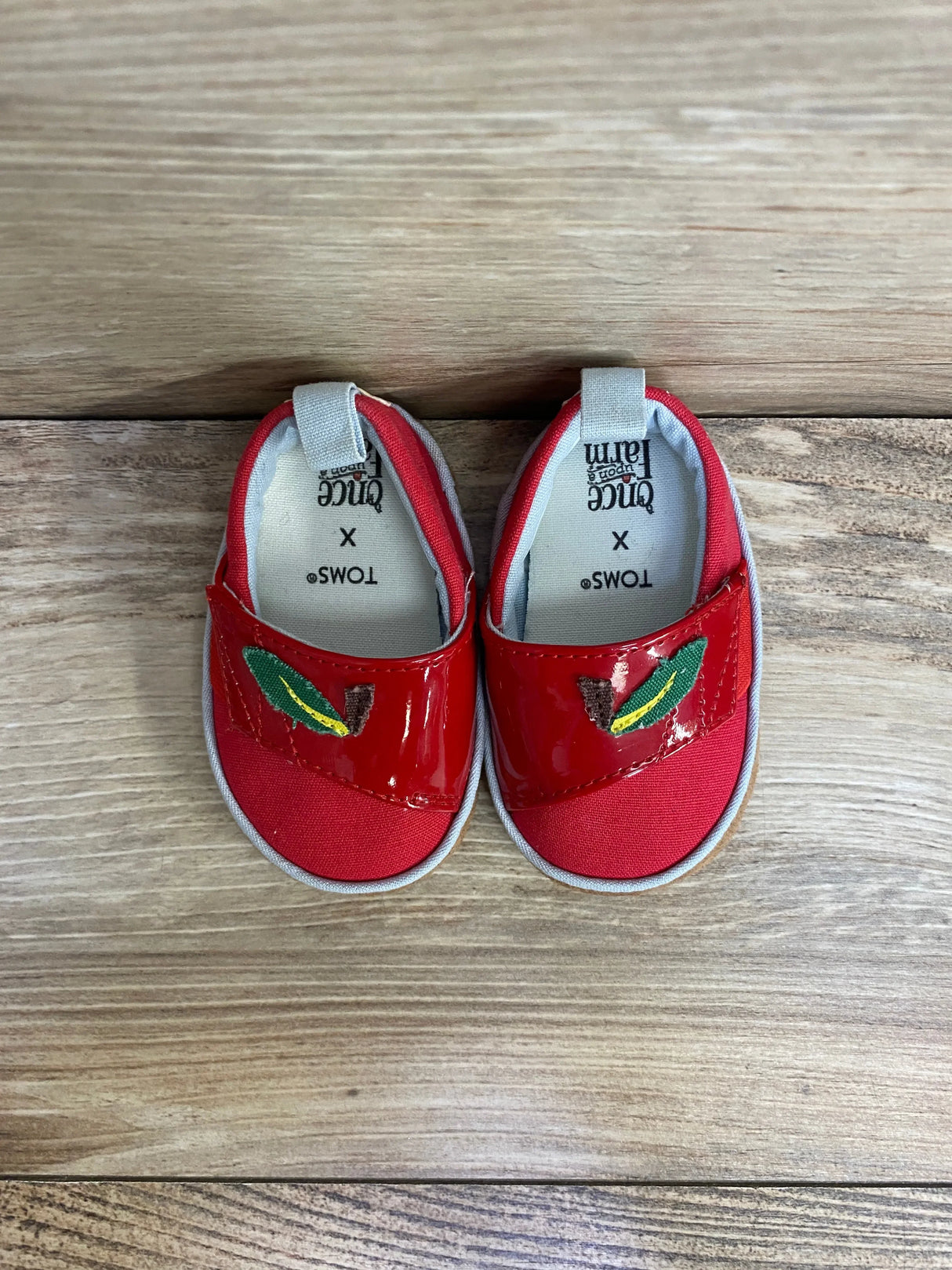 Top-down view of red TOMS Alpargata crib shoes showing glossy patent strap with green embroidered leaf, on light wood floor