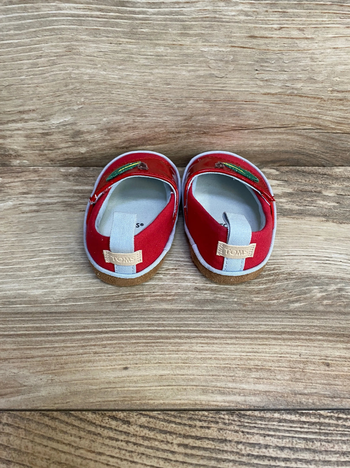 Top view of TOMS Alpargata crib shoes in red, size 3C, showing apple embroidery and elastic heel loop on wood background