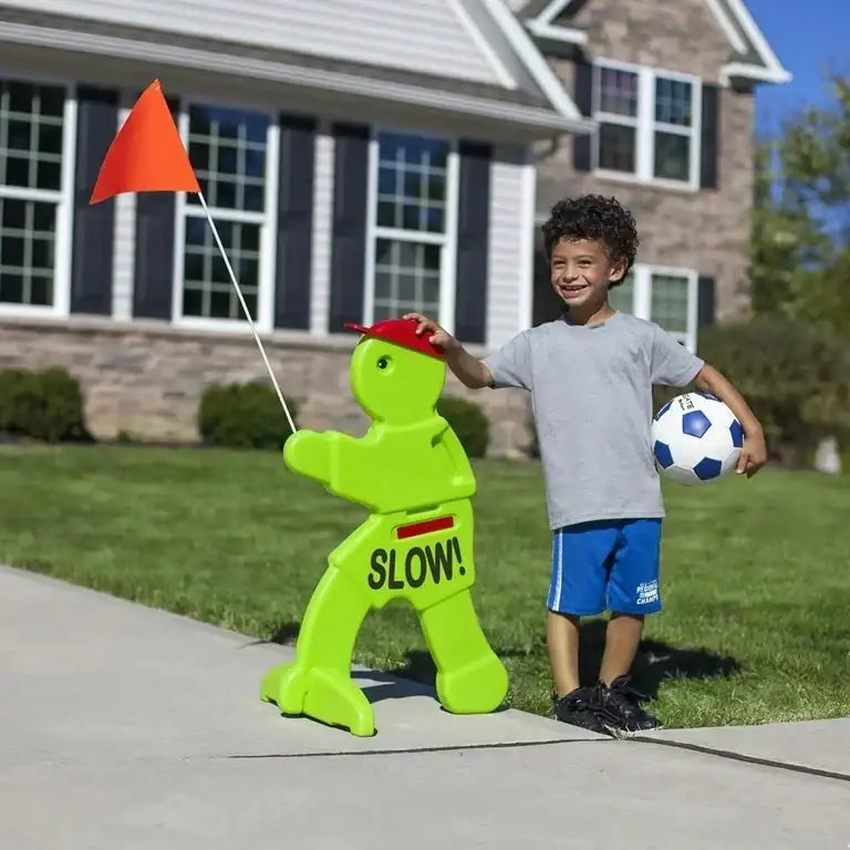 Smiling boy in gray shirt and blue shorts holding soccer ball next to green slow sign with orange flag on sidewalk