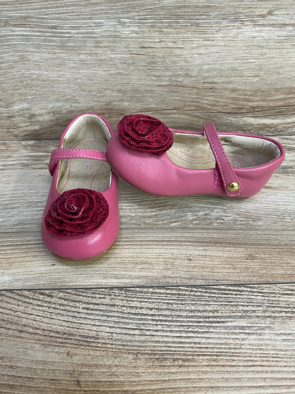 Angled front view of pink Mary Jane toddler shoes size 6-6.5 C showing strap snap and layered rose decoration on wood