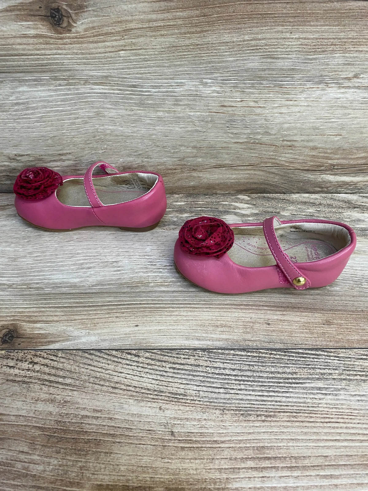 Side view of Pediped Mary Jane toddler shoes showcasing pink leather upper with Velcro strap and dark pink rosette on a wood plank background
