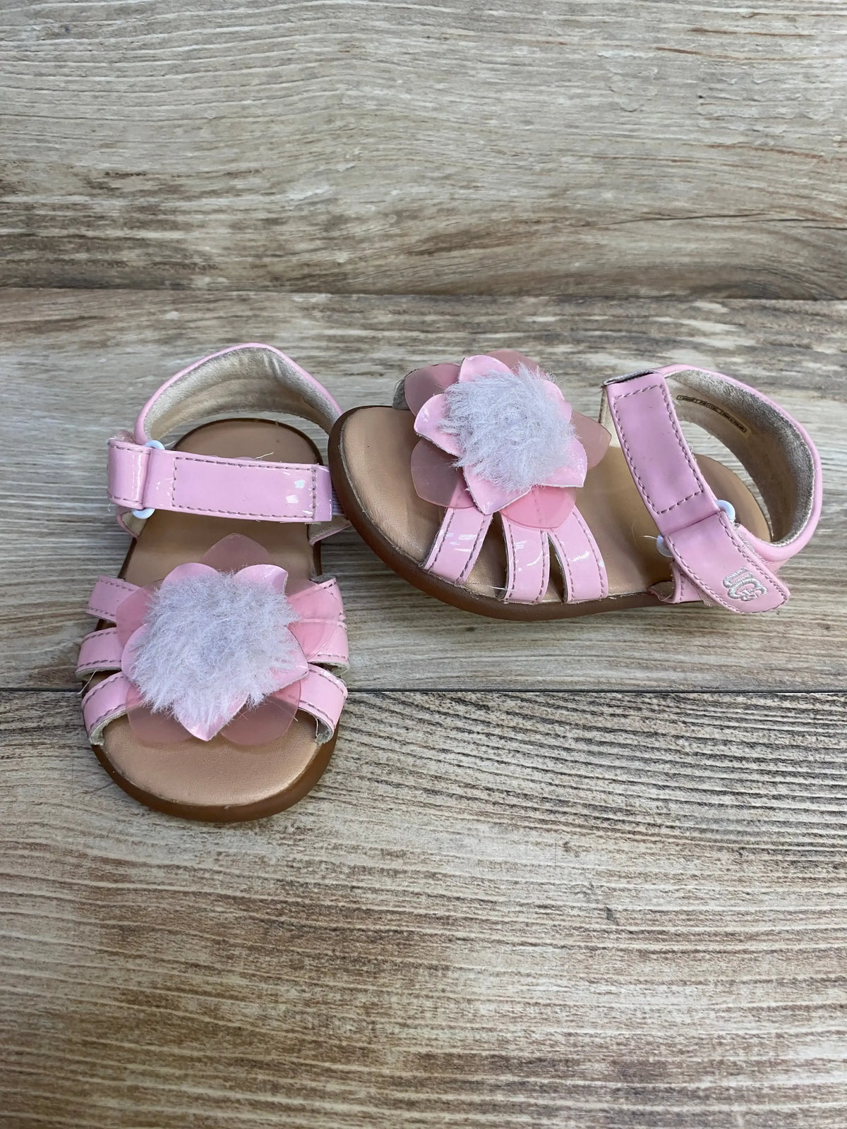 Front and side view of toddler UGG cactus-flower sandals in pink showing fuzzy flower embellishments and velcro ankle straps on wood background