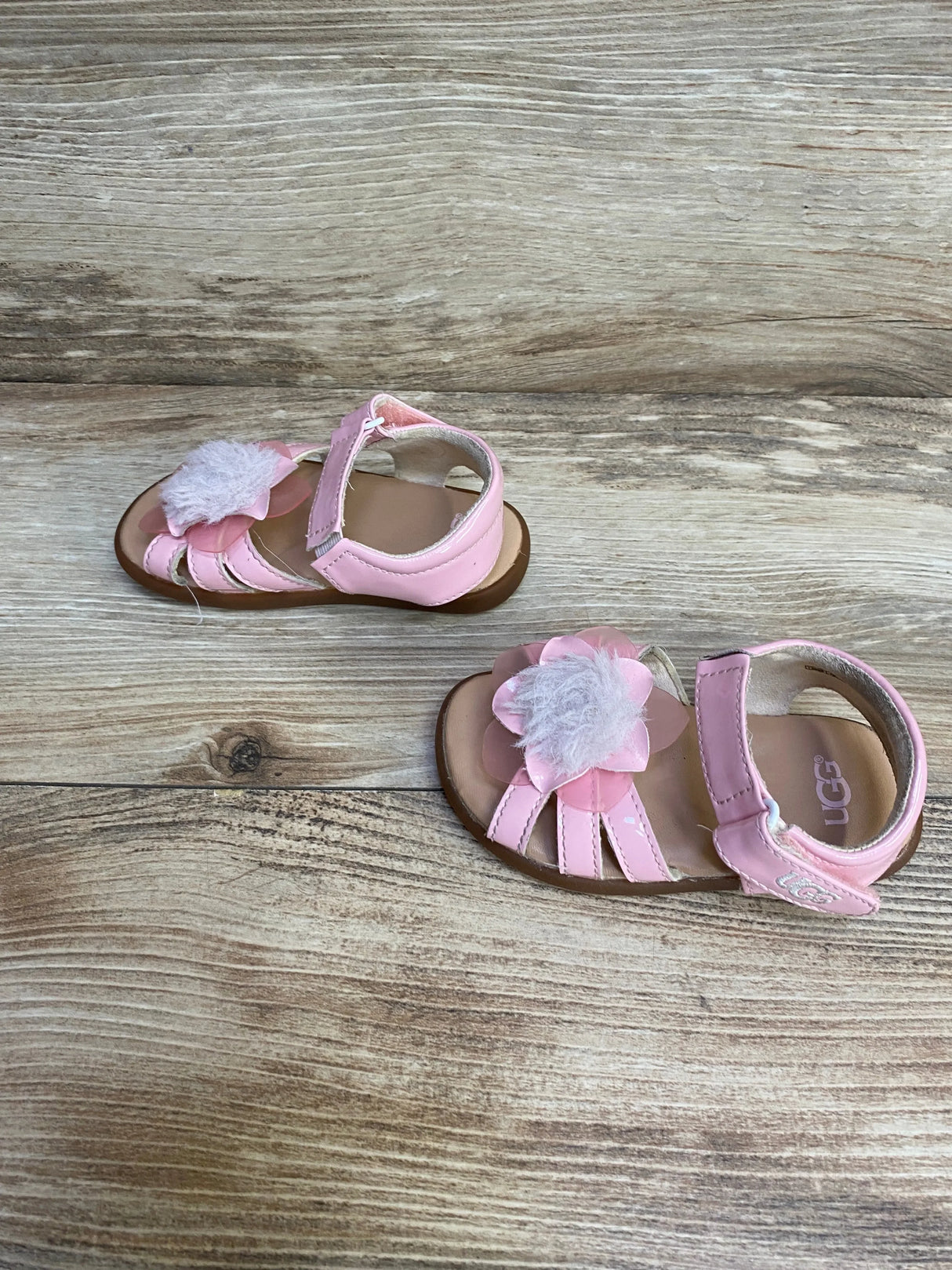 Top view of UGG pink Cactus Flower toddler sandals showing fuzzy flower and Velcro ankle strap on wood background