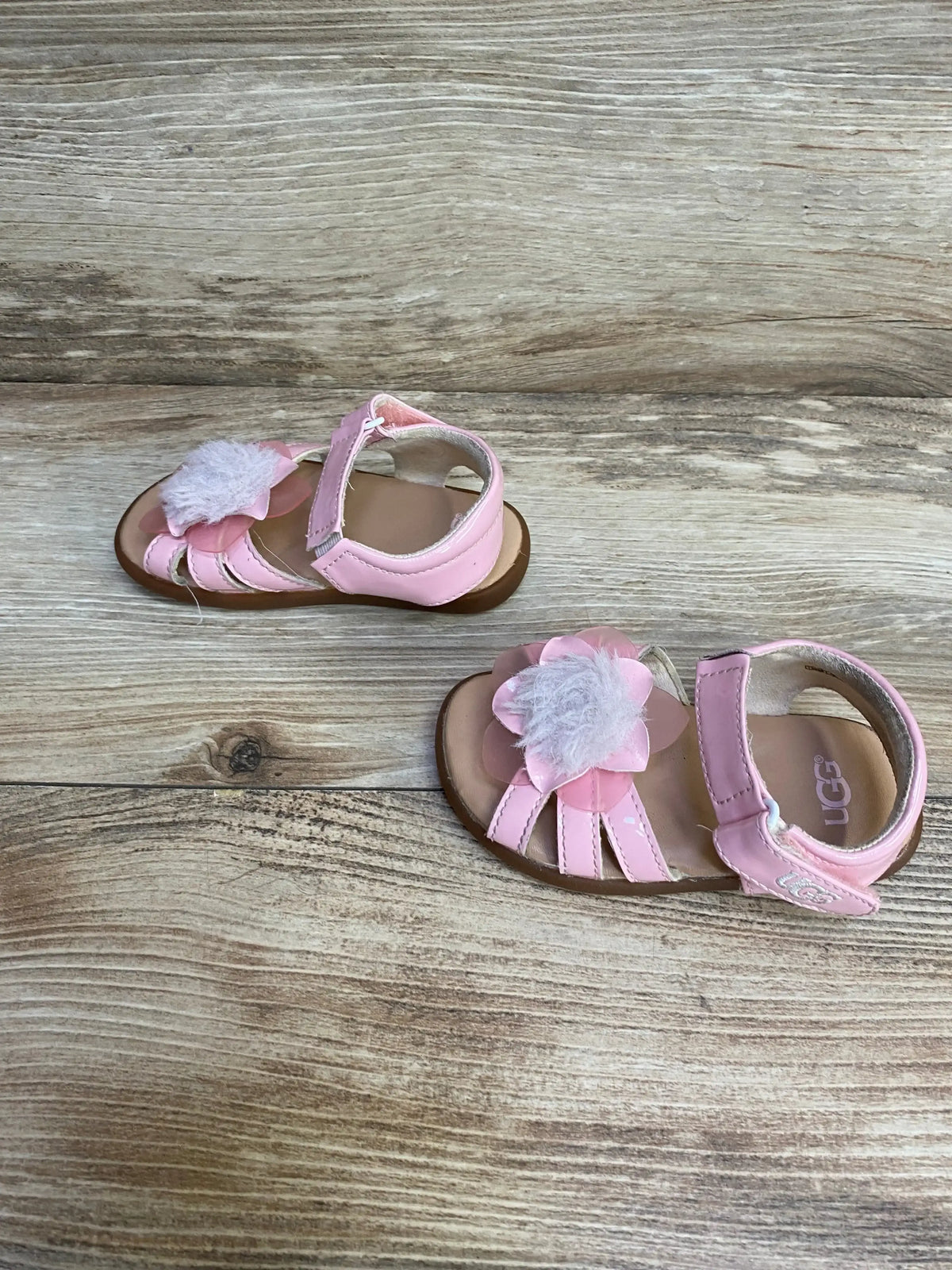 Top view of UGG pink sandals showcasing white tulle flower applique ankle strap and open toe on wood plank floor