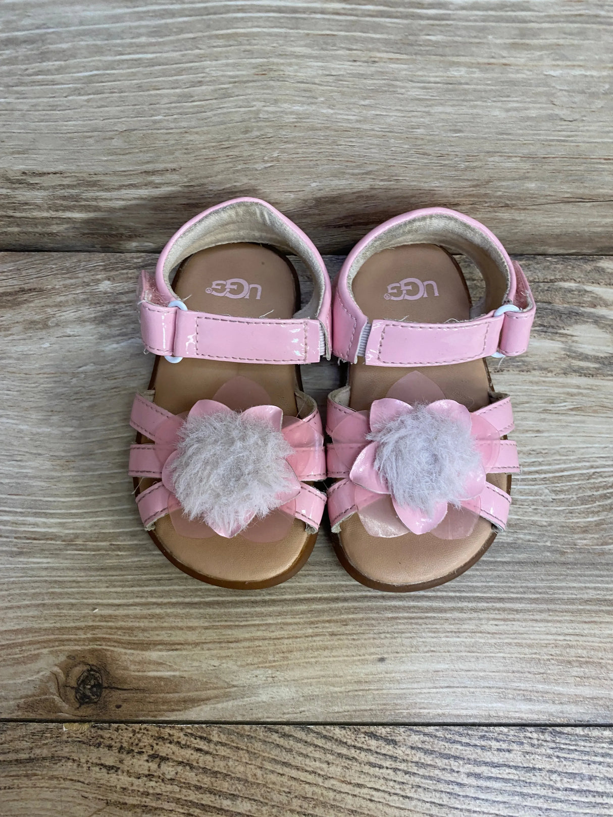Top-down view of UGG child pink patent sandals with flower petals, fuzzy pom-pom and Velcro ankle strap on light wood