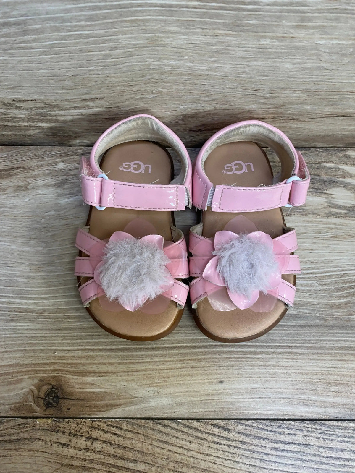 Top-down view of UGG child pink patent sandals with flower petals, fuzzy pom-pom and Velcro ankle strap on light wood