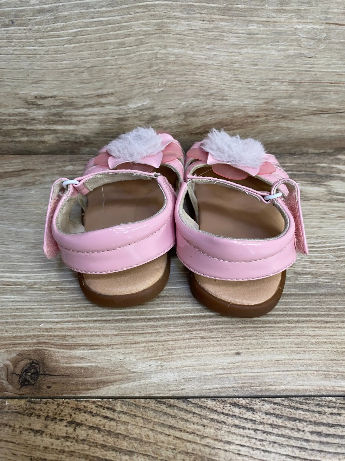 Rear view of pink UGG Cactus Flower sandals showing patent leather flowers, faux fur poms, and hook and loop straps on wood