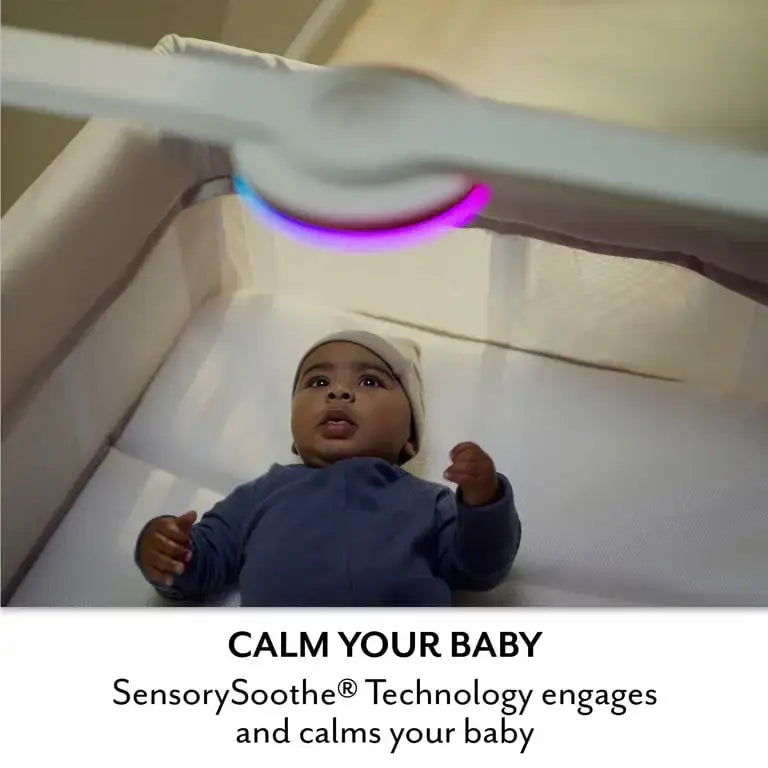 Baby lies in Evenflo Eris playard bassinet under colorful light; Calm your baby with SensorySoothe Technology