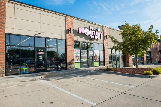 Me n Mommy To Be storefront with sign, glass display windows, parking spaces and a tree in front on a sunny day