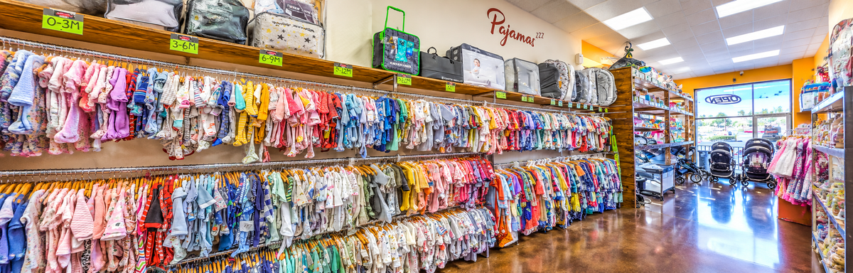 A wide view of a retail store aisle filled with racks of colorful baby clothing and accessories. The store has bright ceiling lights, wooden floors, and shelves displaying strollers and other baby items.