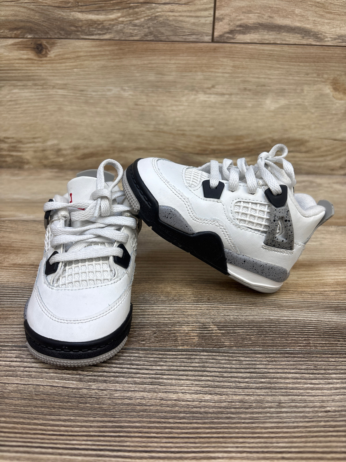 Angled view of Air Jordan 4 Retro OG TD White Cement sneakers showcasing white leather and black detailing on a wood grain surface