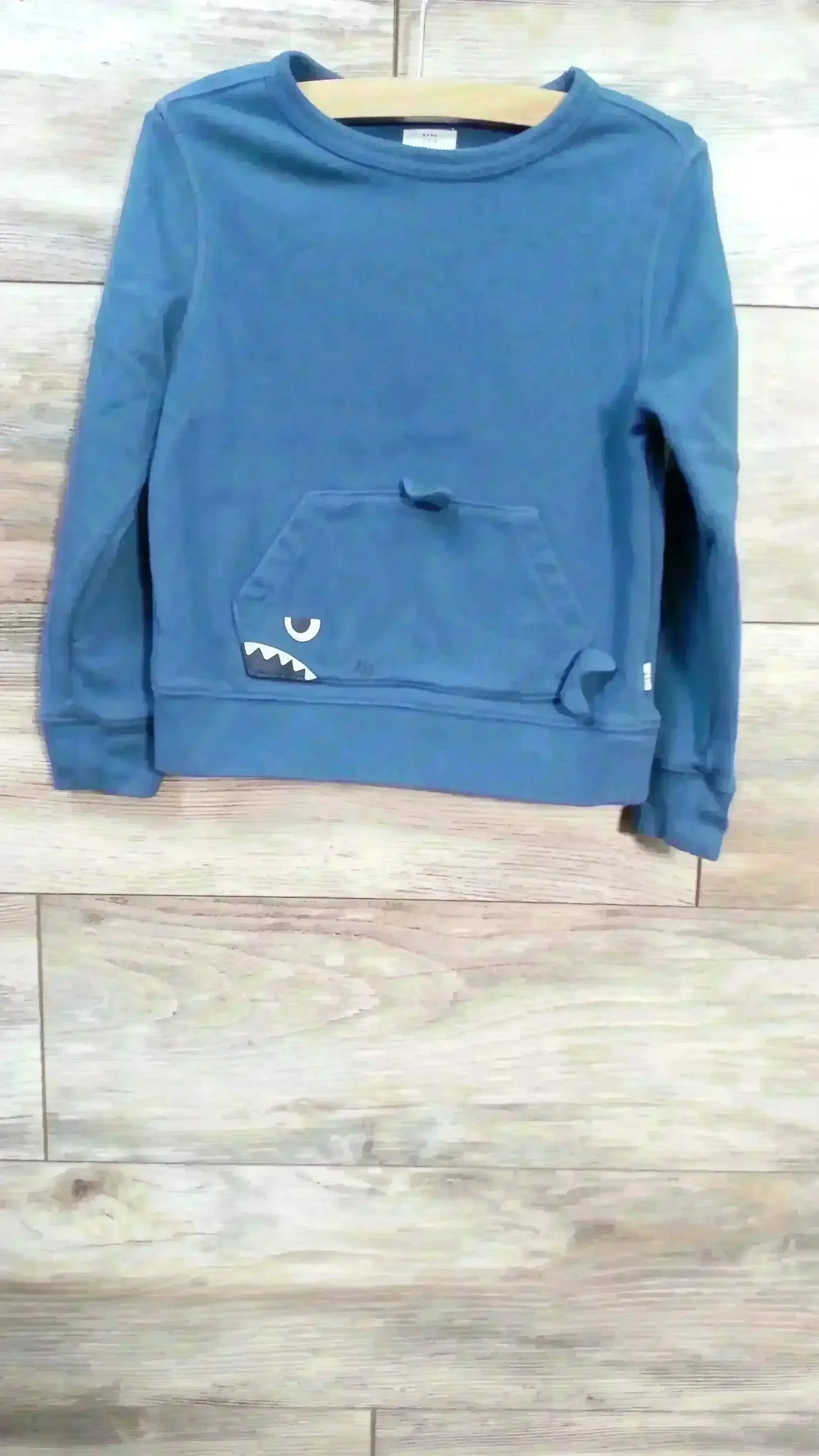 Front of Baby Gap blue sweatshirt (5T) showing shark pocket with printed eye, teeth and fabric fins on light wood floor
