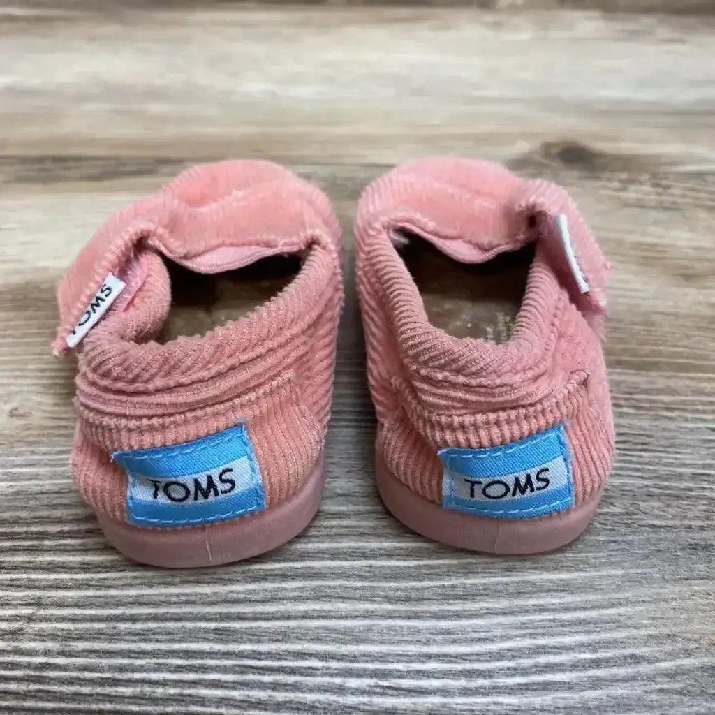 Rear view of pink corduroy Toms baby slip-on shoes size 3C showing back logo tags on a wood floor