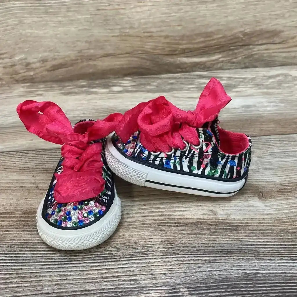 Angled front view of pink Converse low-top sneakers size 2C showing zebra print rhinestone toes and pink ribbon laces on wood