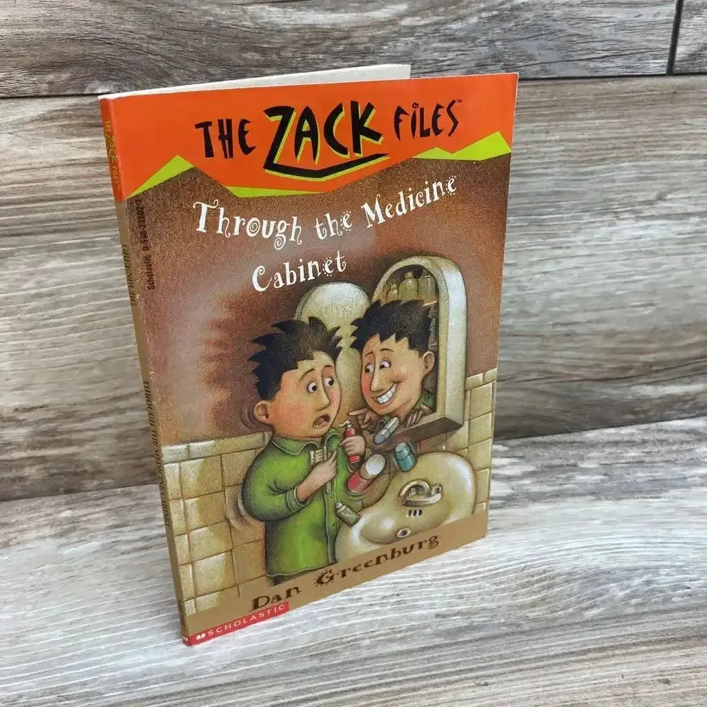 Three-quarter view of paperback Through the Medicine Cabinet (The Zack Files) showing illustrated front cover and spine on wood plank background