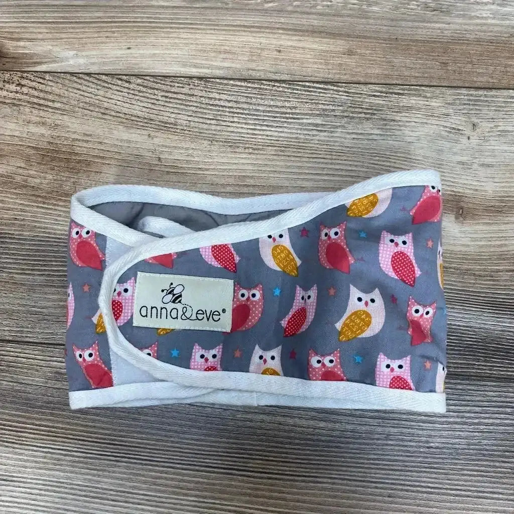 Top view of Anna and Eve swaddle strap, gray with pink and yellow owl print, white trim and visible logo on wood background