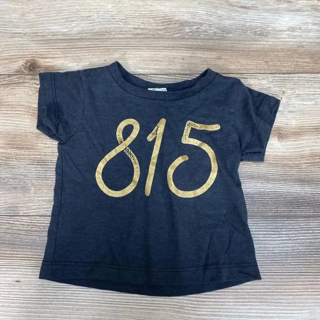 Flat lay of grey toddler short-sleeve shirt size 6-12 months showcasing large gold 815 graphic on front with light wood background