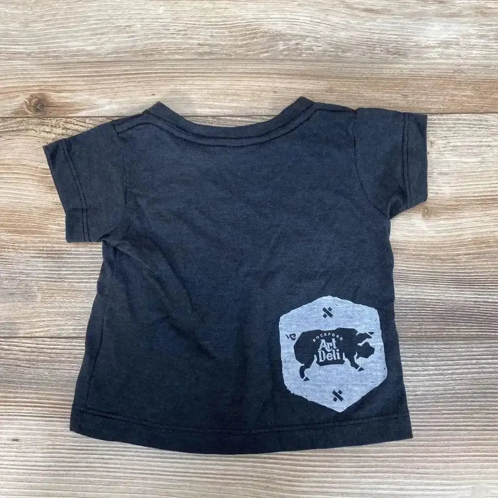 Flat lay of grey 6-12m baby t-shirt showing lower-right Art Deli cow graphic on wood background
