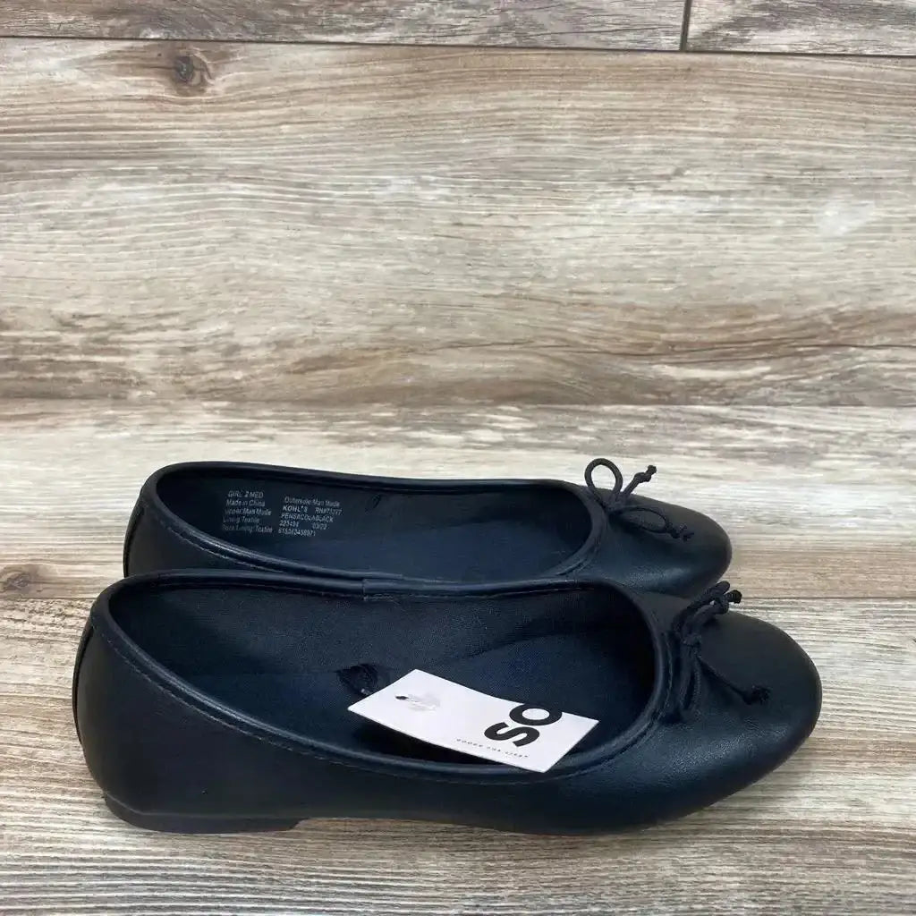 Three-quarter view of NWT SO Pensacola Ballet Flats size 2y showcasing black faux leather with bow accents and brand hangtag on wood plank floor
