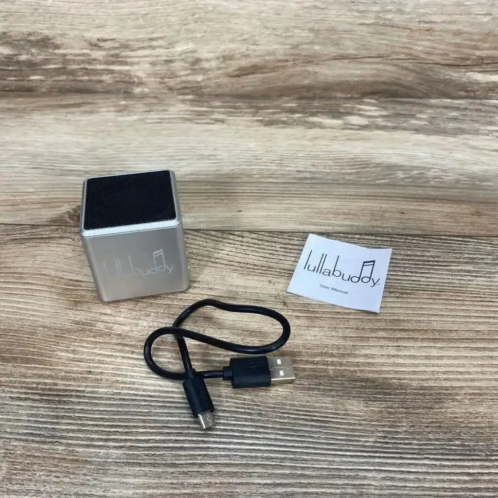 Three-quarter view of grey Lullabuddy portable speaker with black grill, logo, micro USB cable and manual on wood background