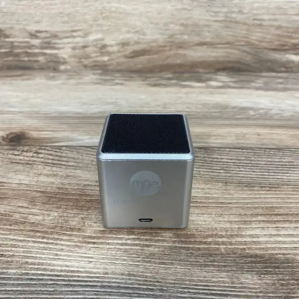 Front view of grey Lullabuddy portable speaker showing cube metal body, black top grille and charging port on a wood tabletop