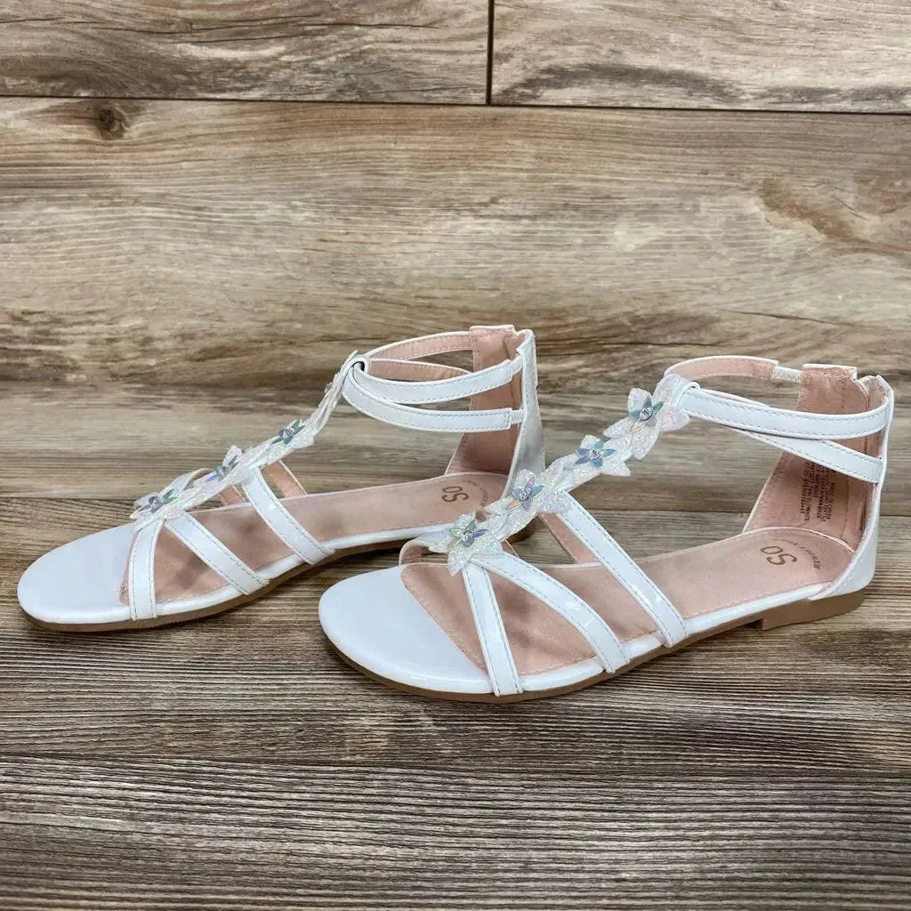 Side view of a pair of white toddler gladiator sandals showing flower-embellished T-straps and ankle straps on wood background