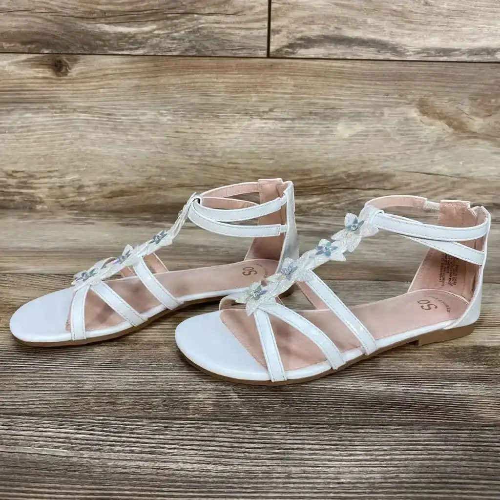 Side view of a pair of white toddler gladiator sandals showing flower-embellished T-straps and ankle straps on wood background