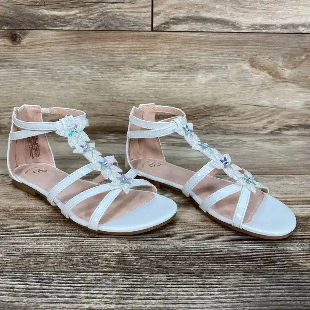 Angled view of white New SO Loganberry gladiator sandals showing iridescent flower accents and ankle straps on wood floor