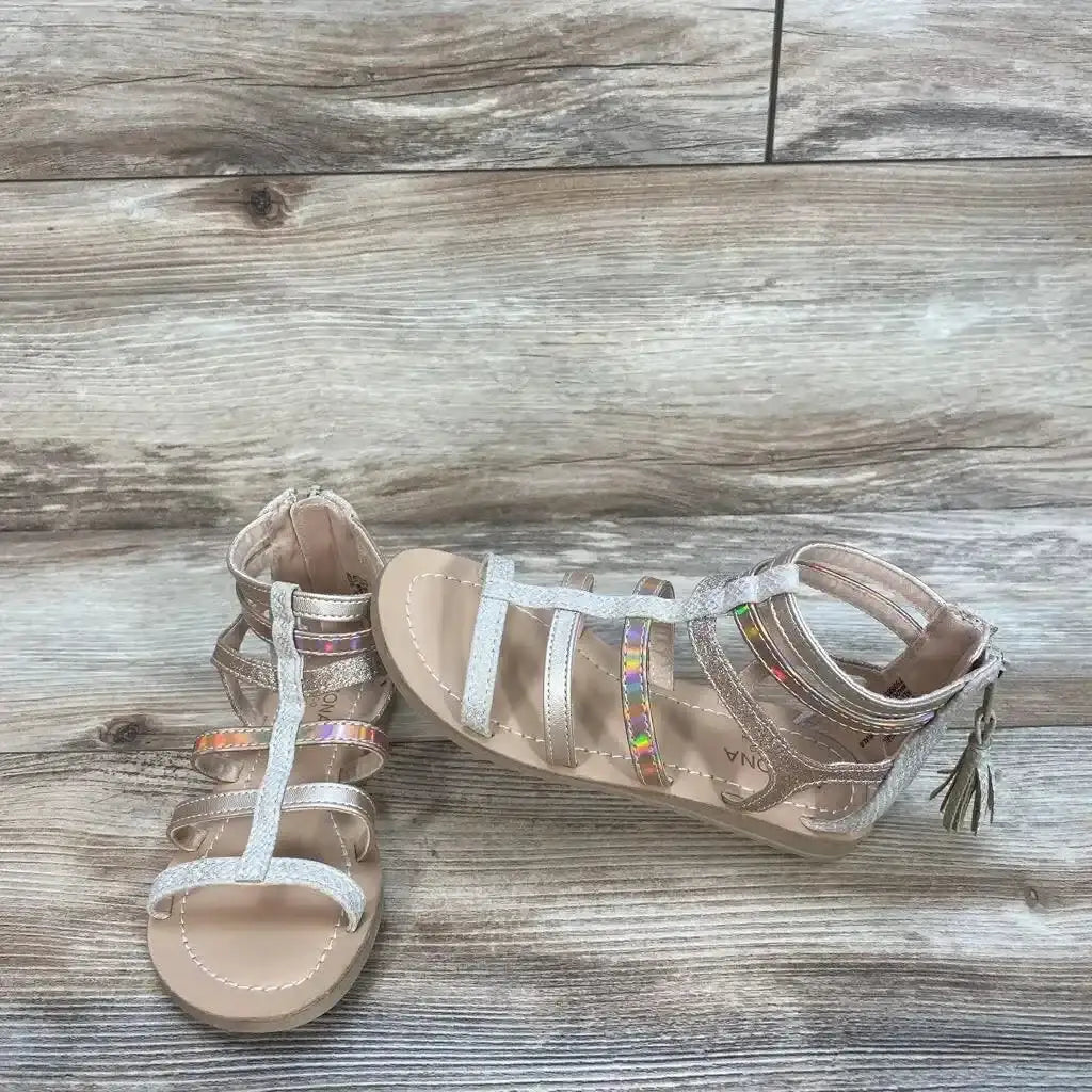 Three quarter view of Arizona Jean Co Angel gladiator sandals showcasing rose gold straps with iridescent accents and hangtag on wood plank floor