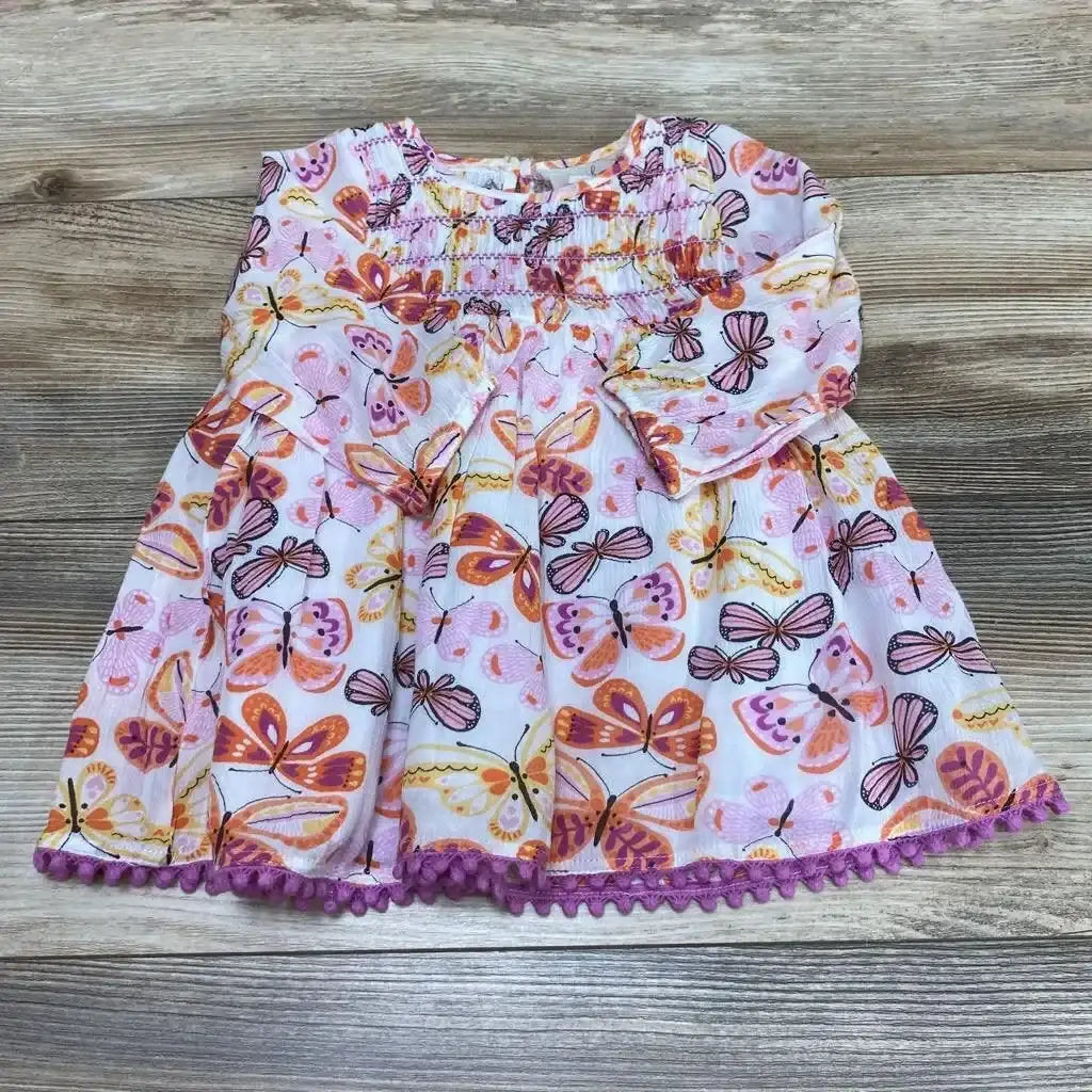 Flat lay of pink smocked baby dress (6-12m) showing butterfly all-over print, long sleeves and purple pom-pom trim on wood background