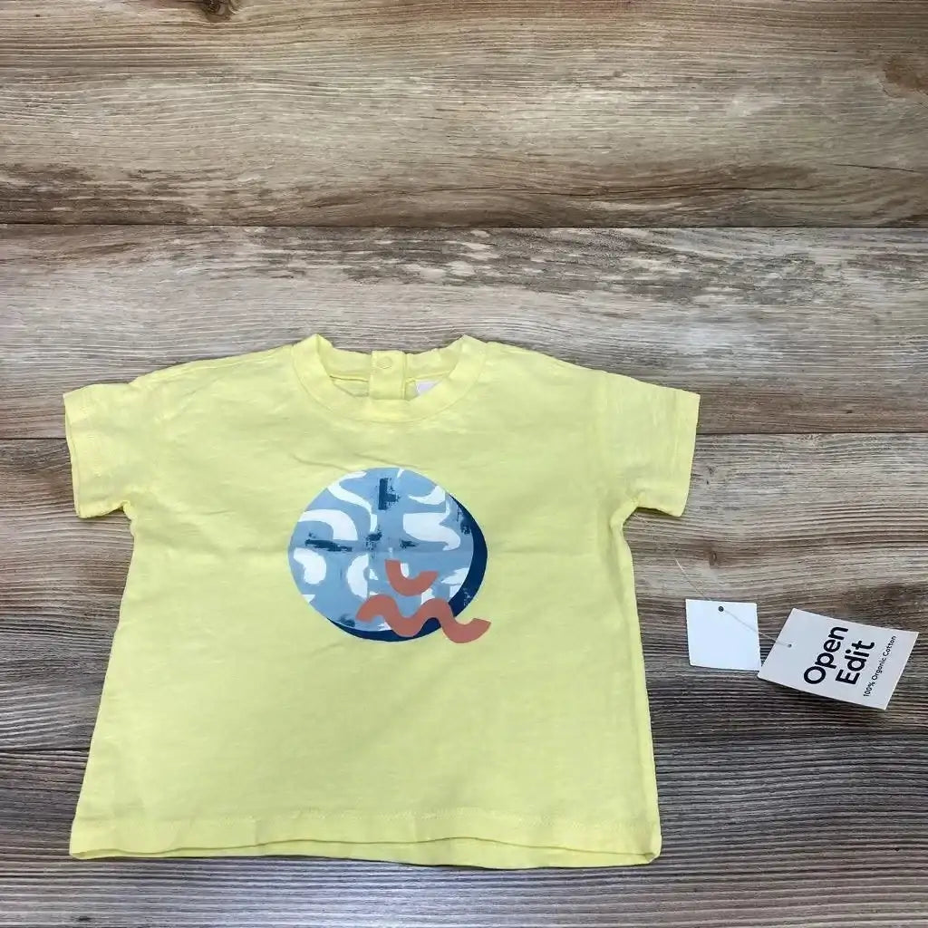 Flat lay of yellow Open Edit organic baby t-shirt size 12m showing round blue graphic and attached tag on wood background