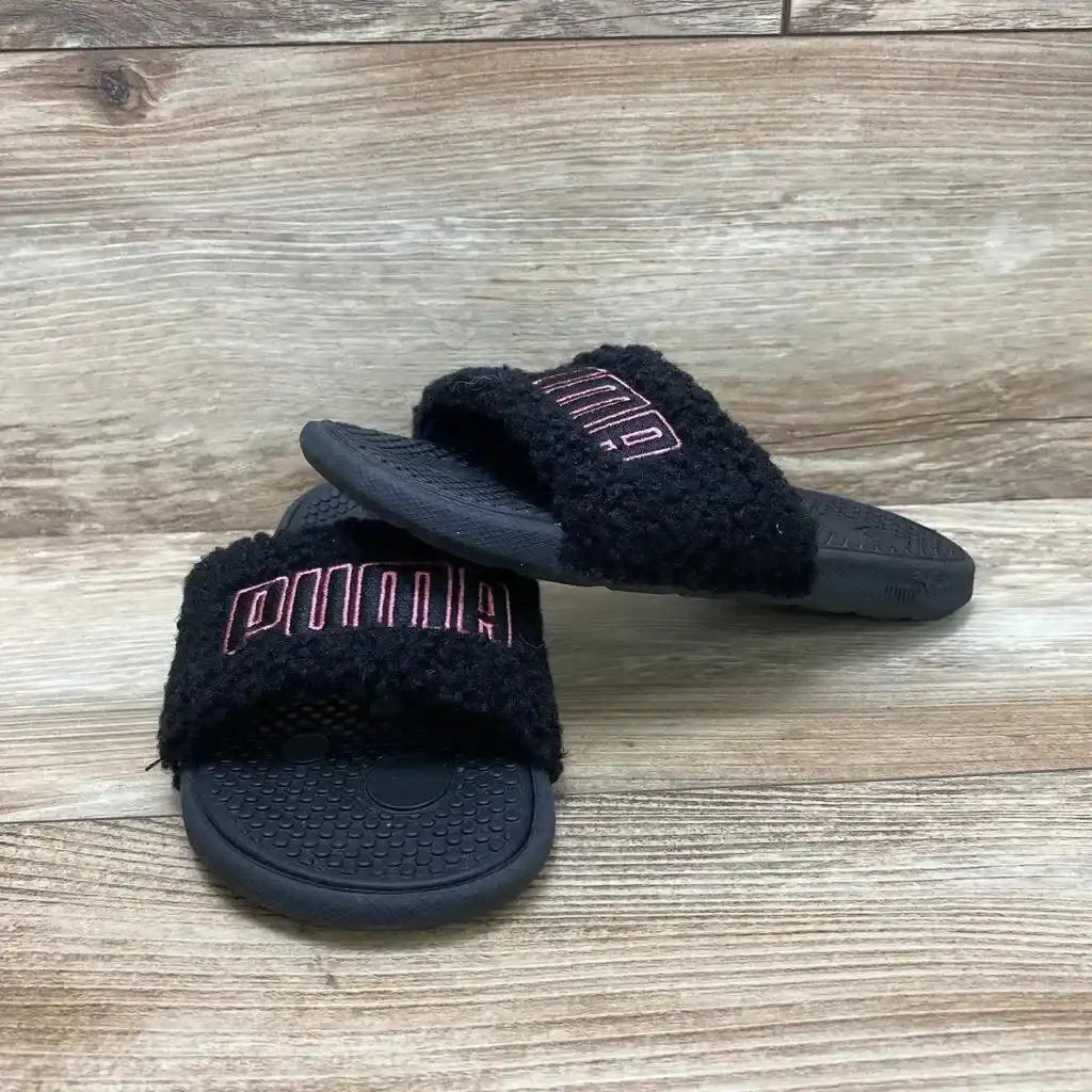 Angled front view of black Puma sherpa slides showcasing fuzzy strap with pink logo and textured footbed on wood background