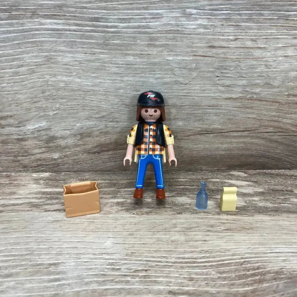 Front view of Playmobil 5665 dump truck driver figure with black helmet, plaid shirt, blue pants and three small accessories on wood background