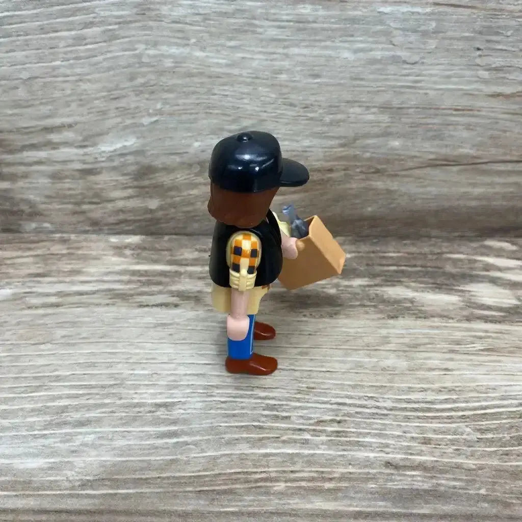 Side view of Playmobil 5665 dump truck driver figure holding a small brown box of accessories on a wood-plank background