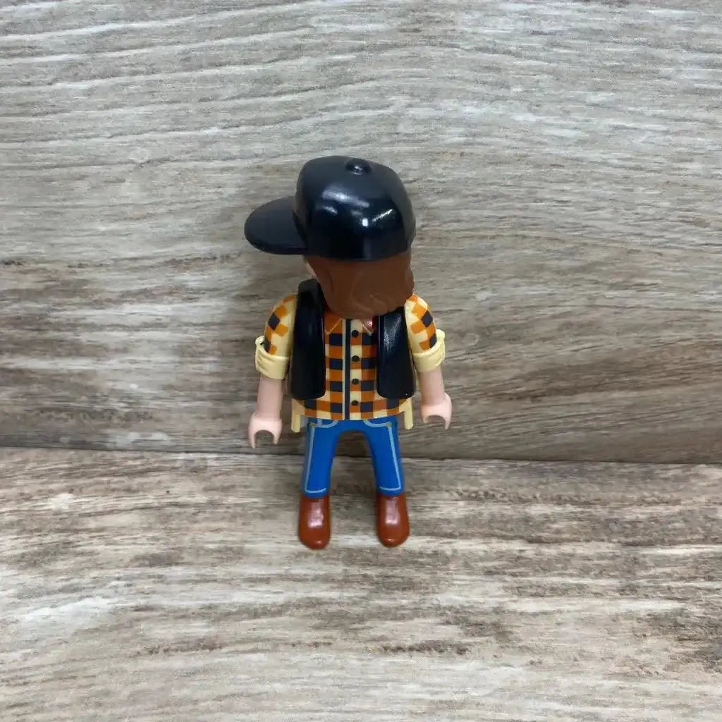 Rear view of Playmobil 5665 dump truck driver figure showcasing black cap, orange checkered shirt, black vest and blue pants on wood background