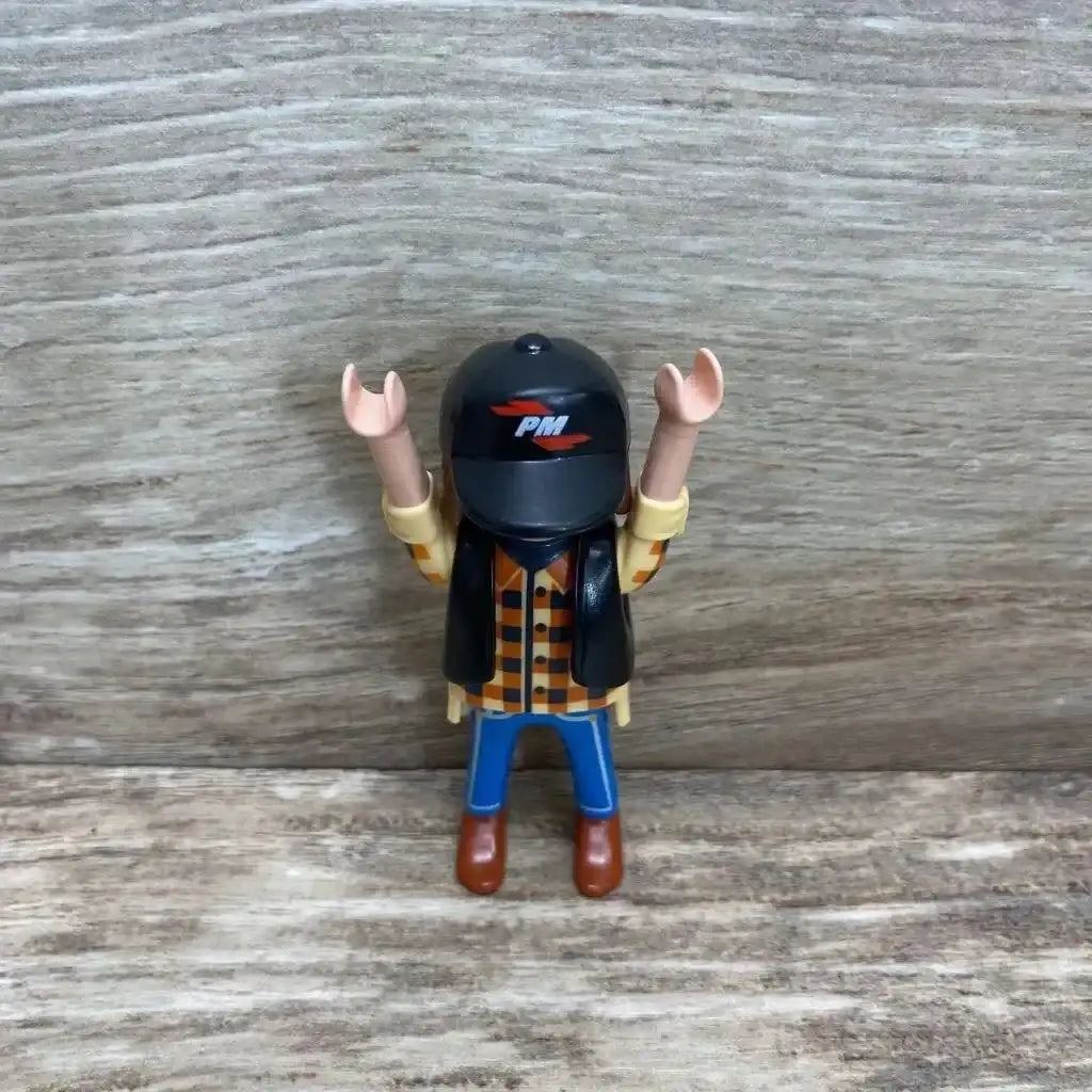 Front view of Playmobil 5665 dump truck driver figure with black helmet showing PM logo, black vest, orange checked shirt, blue pants on wood background