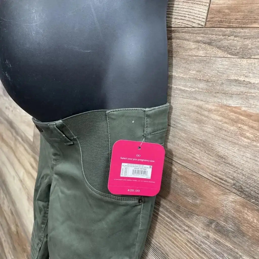 Side view of olive maternity utility jeggings showing elastic belly panel, cargo pocket, and pink tag on wood background