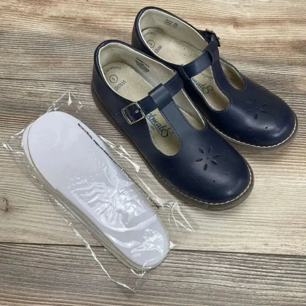 Top-down view of navy Sherry Mary Jane shoes with T-strap buckle, floral cutouts, and white insoles in plastic on wood floor