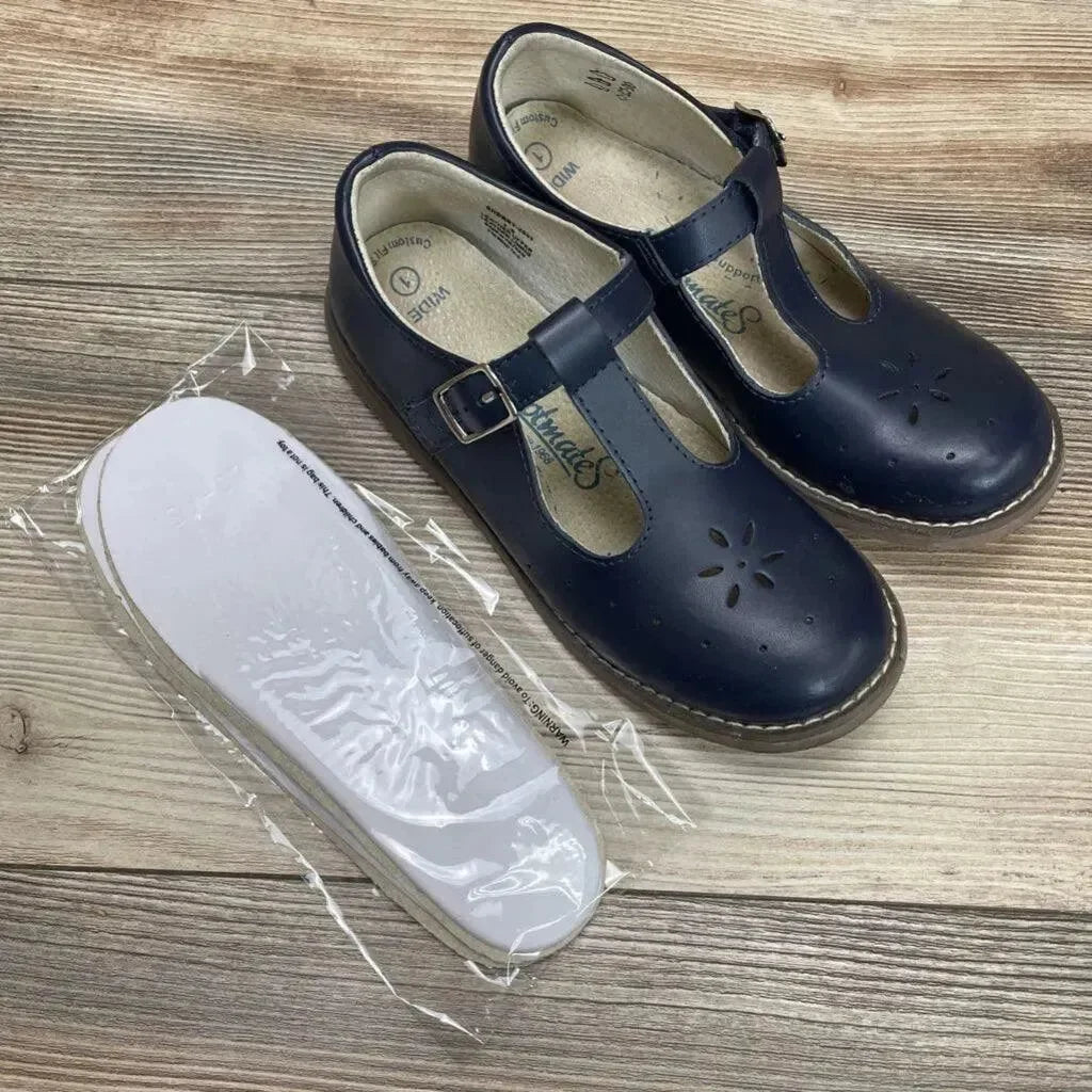 Top-down view of navy Sherry Mary Jane shoes with T-strap buckle, floral cutouts, and white insoles in plastic on wood floor