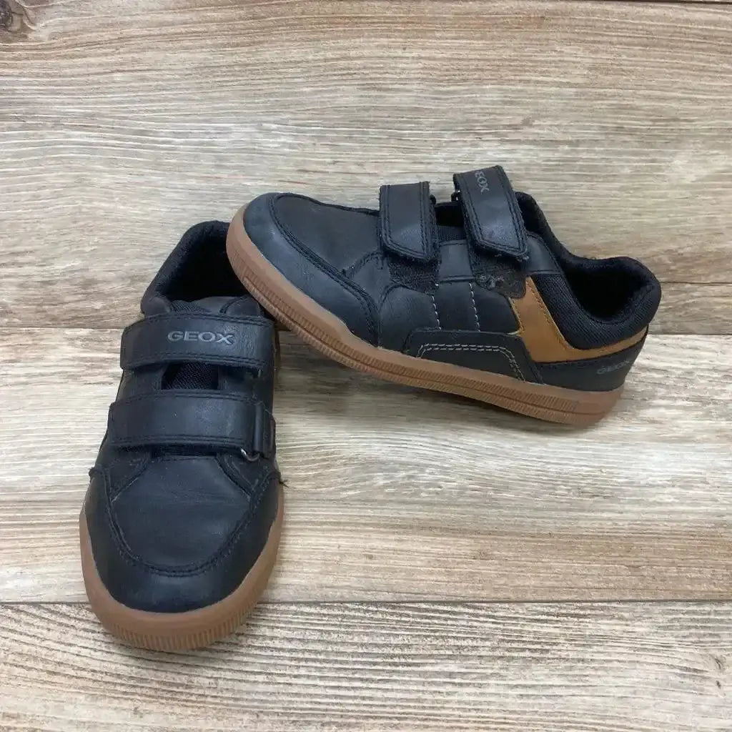 Front three quarter view of black Geox Arzachboy toddler sneakers with Velcro straps and brown gum soles on wood background