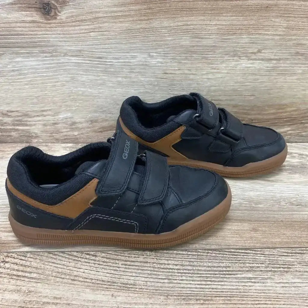 Side view of black Geox Arzachboy toddler sneakers (size 13c) with brown heel accents and two hook-and-loop straps on a wood floor background