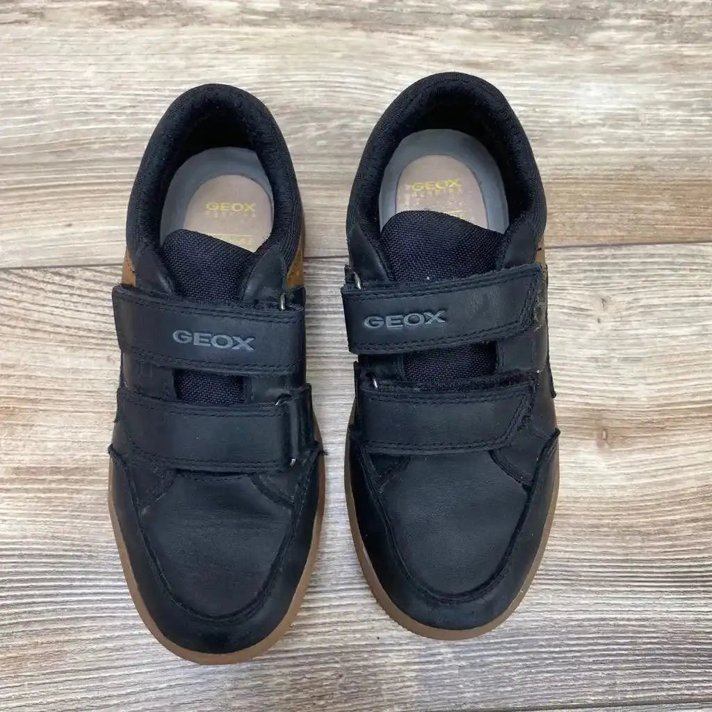 Top-down view of black Geox kids' sneakers showing double velcro straps and gum rubber soles on a light wood floor