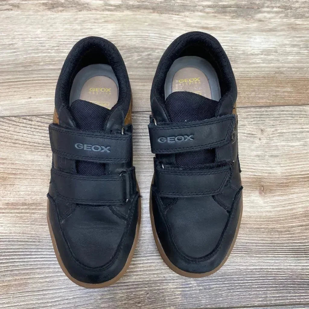 Top-down view of black Geox kids' sneakers showing double velcro straps and gum rubber soles on a light wood floor